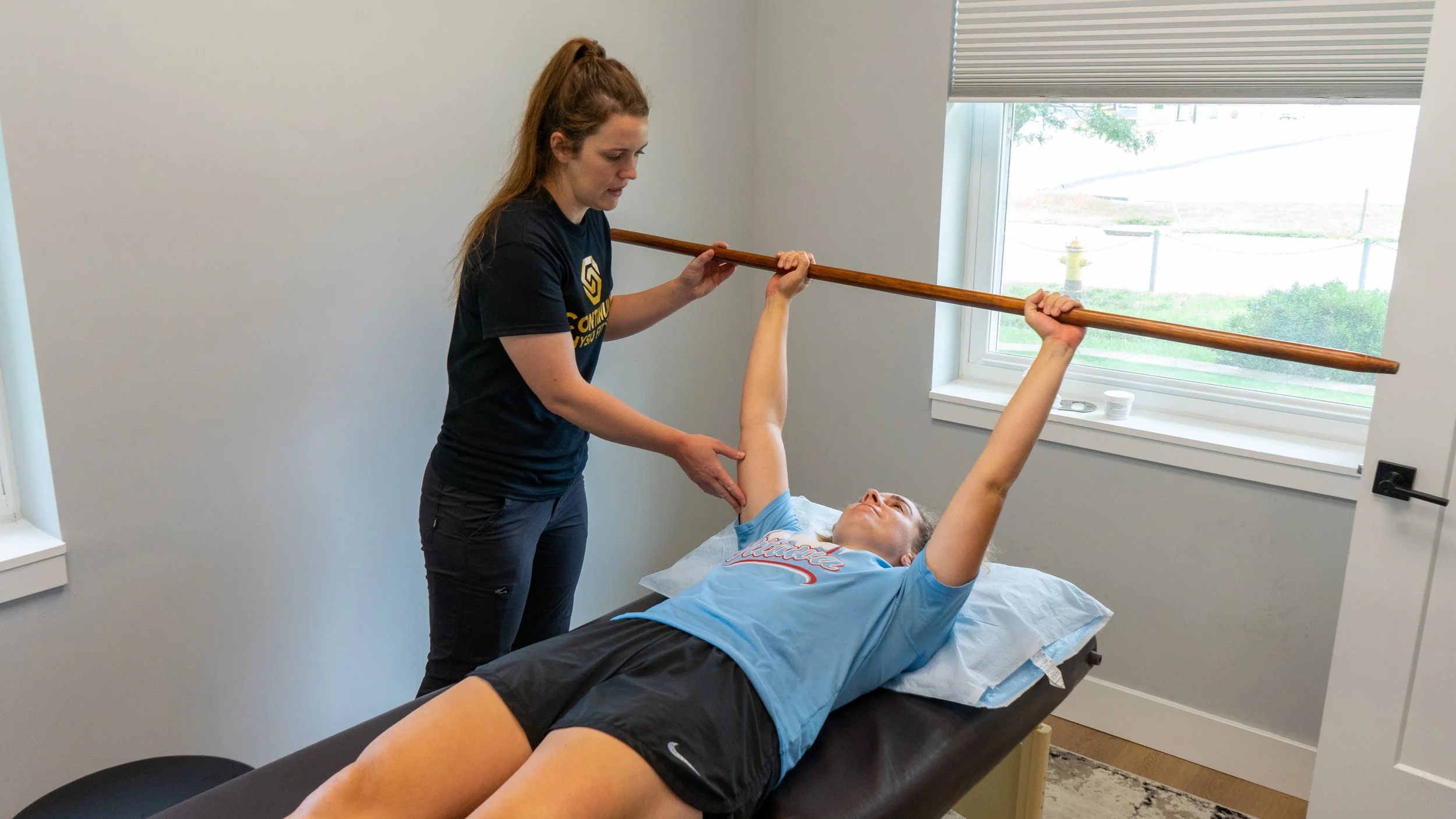 Therapist helping a young boy perform a physical exercise with a wooden stick while lying on a treatment table in a therapy room with a window.