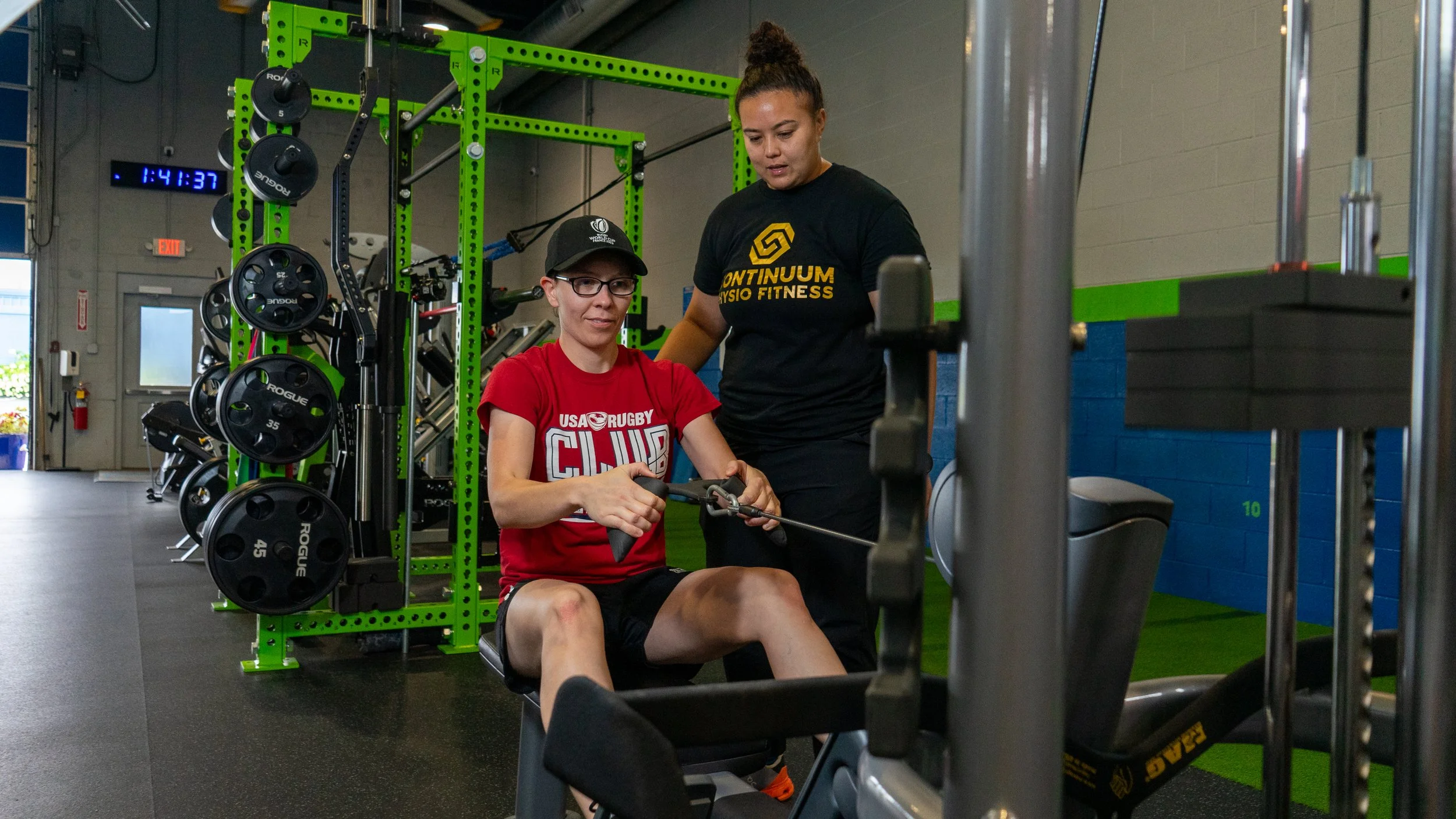 A woman sitting on a rowing machine and a trainer standing next to her, assisting at a gym with weightlifting equipment and a digital clock on the wall.