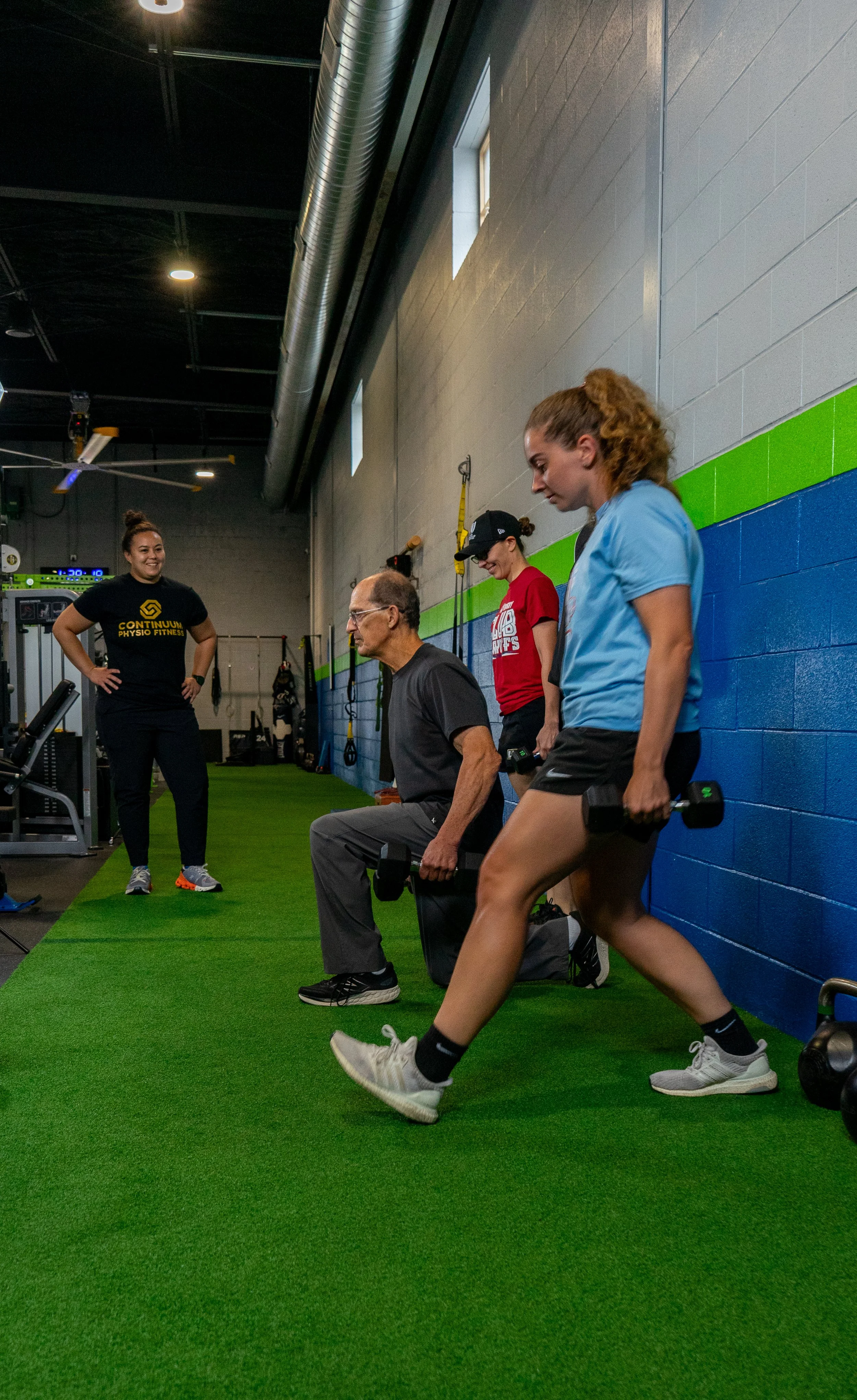 Three women and one man working out in an indoor gym. The man is seated and lifting dumbbells, while two women are aiding with the exercise, and one woman is standing and monitoring. The gym has a green artificial turf floor and a blue wall.