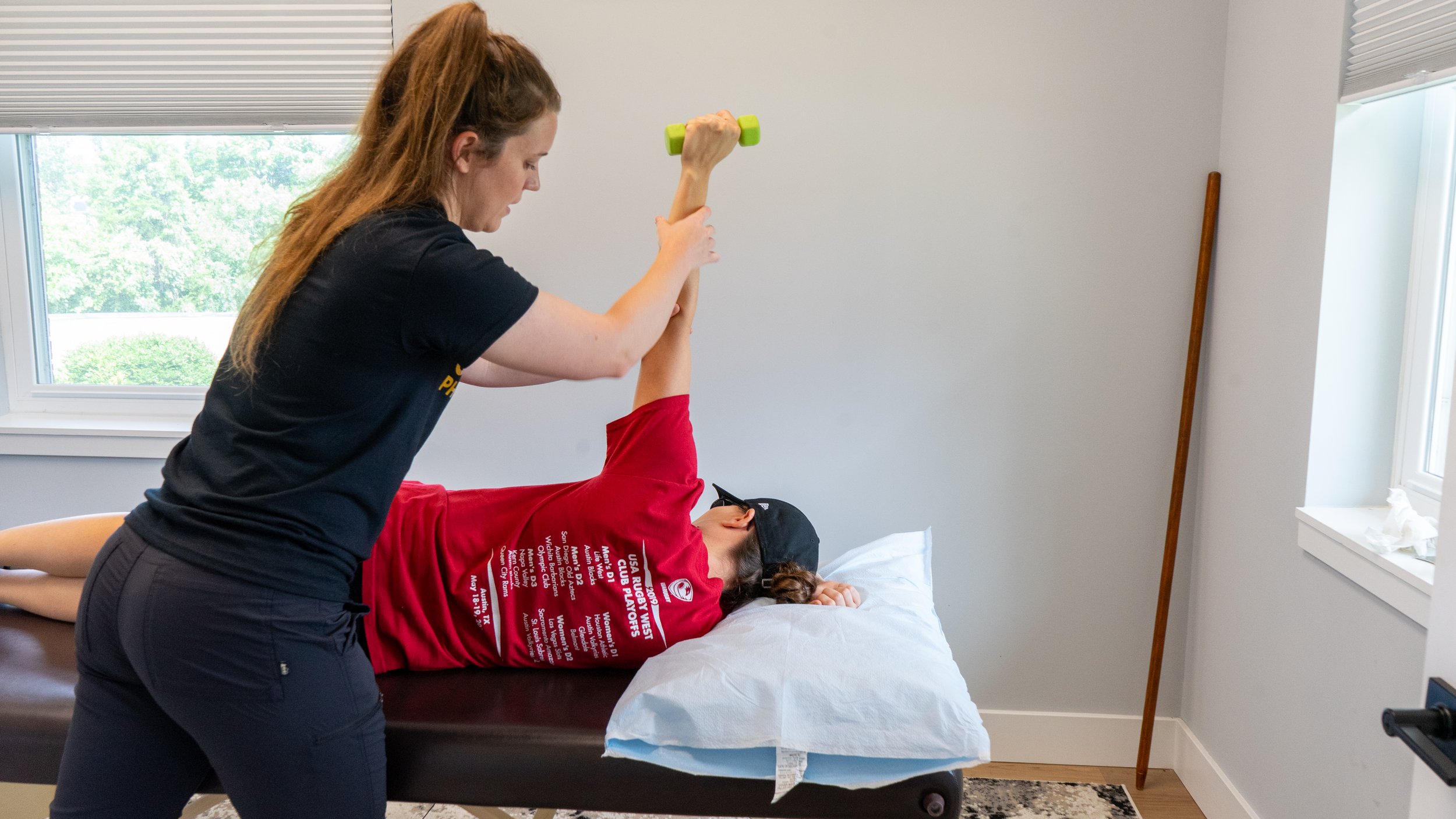 A woman assists a person lying on a bed practicing physical therapy or exercise with a green dumbbell. The person on the bed is wearing a red shirt and a cap. The room has white walls, a window with blinds, and a wooden walking stick leaning against the wall.