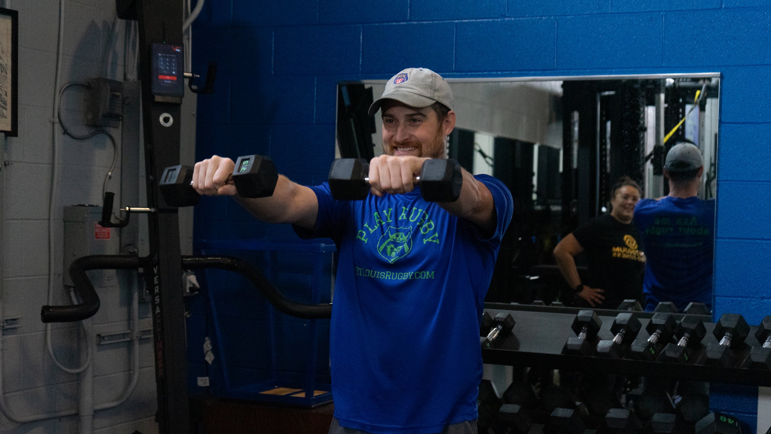 A man with a beard wearing a blue shirt and a gray cap is lifting a dumbbell in a gym. There is a mirror behind him reflecting his image and a woman standing in the background.