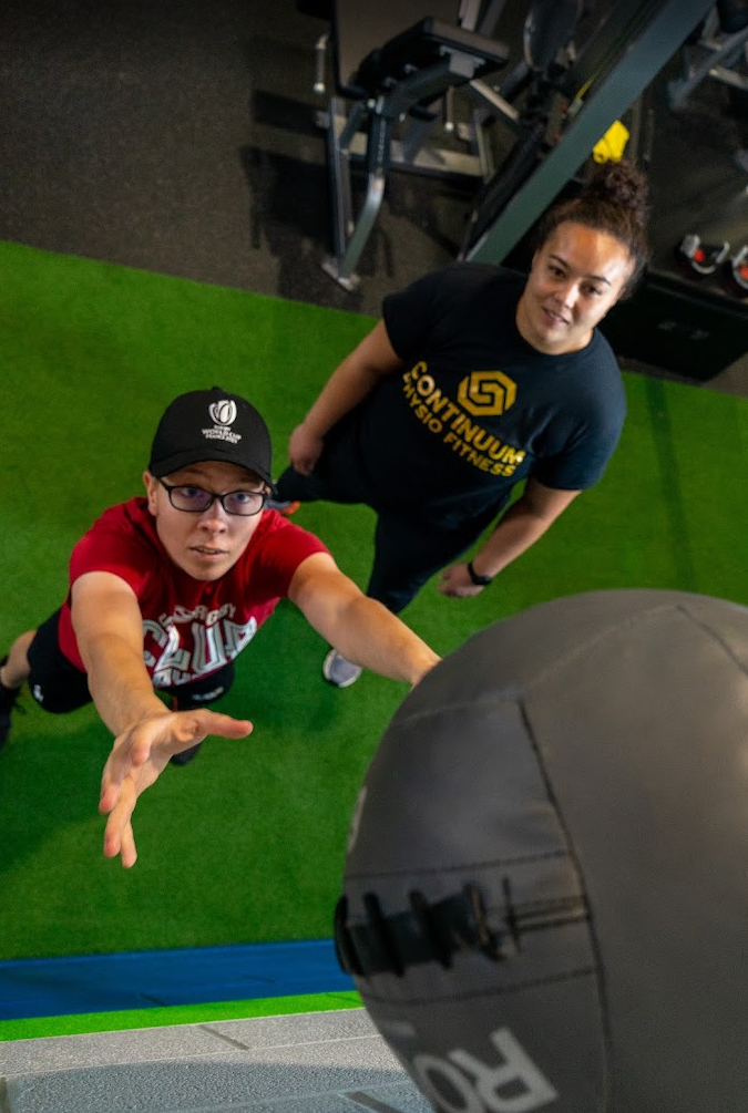 A person in a red shirt and glasses reaches for a medicine ball in a gym, while an instructor observes from behind.