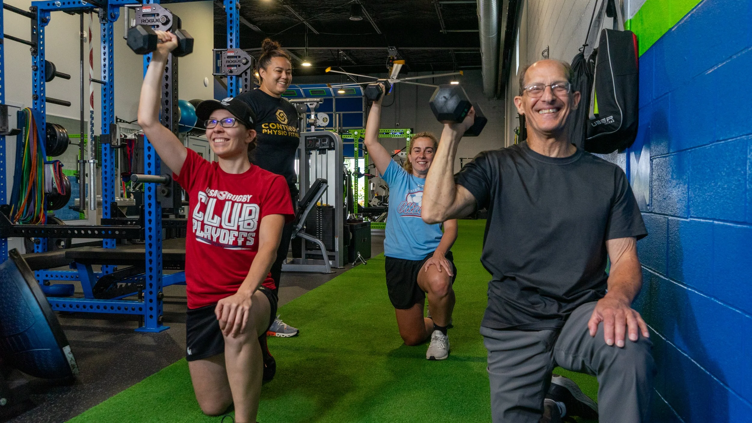 Four people at a gym exercising. Two women are holding dumbbells while kneeling, and a woman and a man behind them are also holding dumbbells overhead. The group appears happy and energetic, with gym equipment visible in the background.
