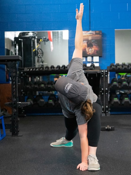 A woman performing a yoga pose in a gym with a blue wall, black rubber flooring, and weight racks in the background.
