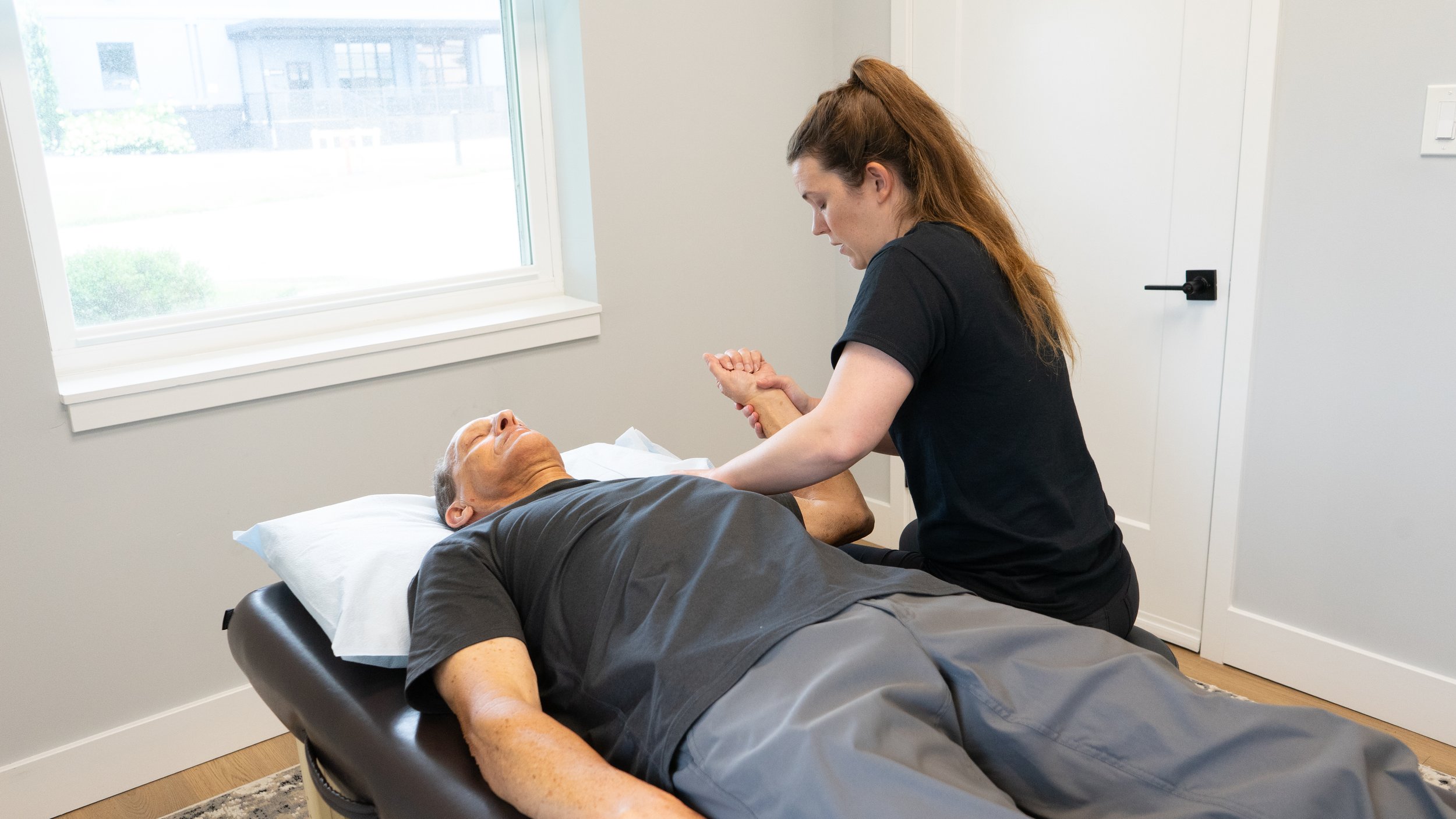 A woman providing physical therapy or massage to a man lying on a treatment table in a medical or wellness room with a window.