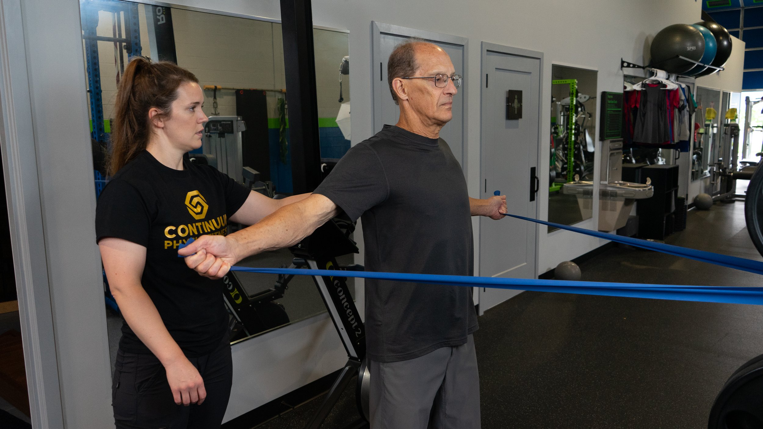 A young woman assisting an older man with a resistance band exercise in a gym. The woman is wearing a black T-shirt with yellow text, and the older man is in a black T-shirt and gray pants. Gym equipment and exercise balls are visible in the background.