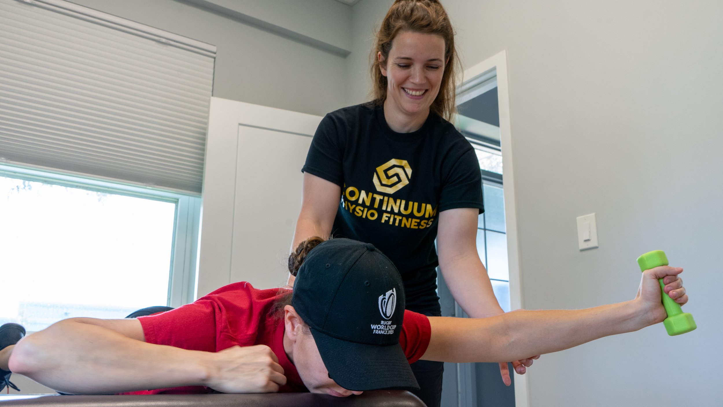 A woman in a black t-shirt with yellow lettering and a logo, smiling, working with a person lying face down on a massage table. The person on the table wears a red shirt and a black cap, and is holding a small green weight in one hand. The room has a window and a white wall with a light switch.