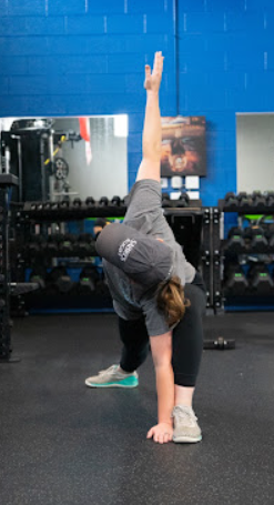 Person stretching or exercising in a gym, touching the floor with one hand, with one arm raised.