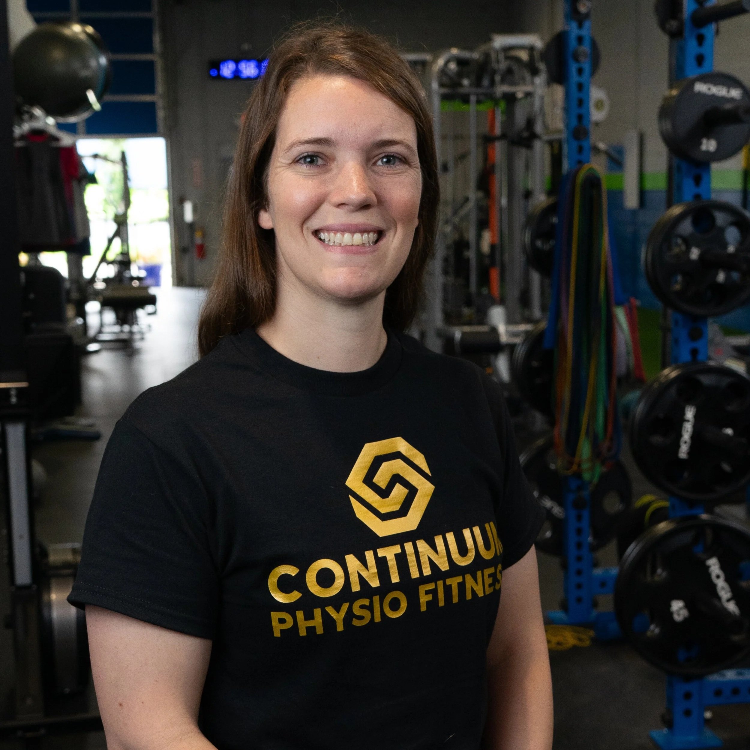 A smiling woman with brown hair wearing a black T-shirt with yellow text, standing in a gym with exercise equipment in the background.