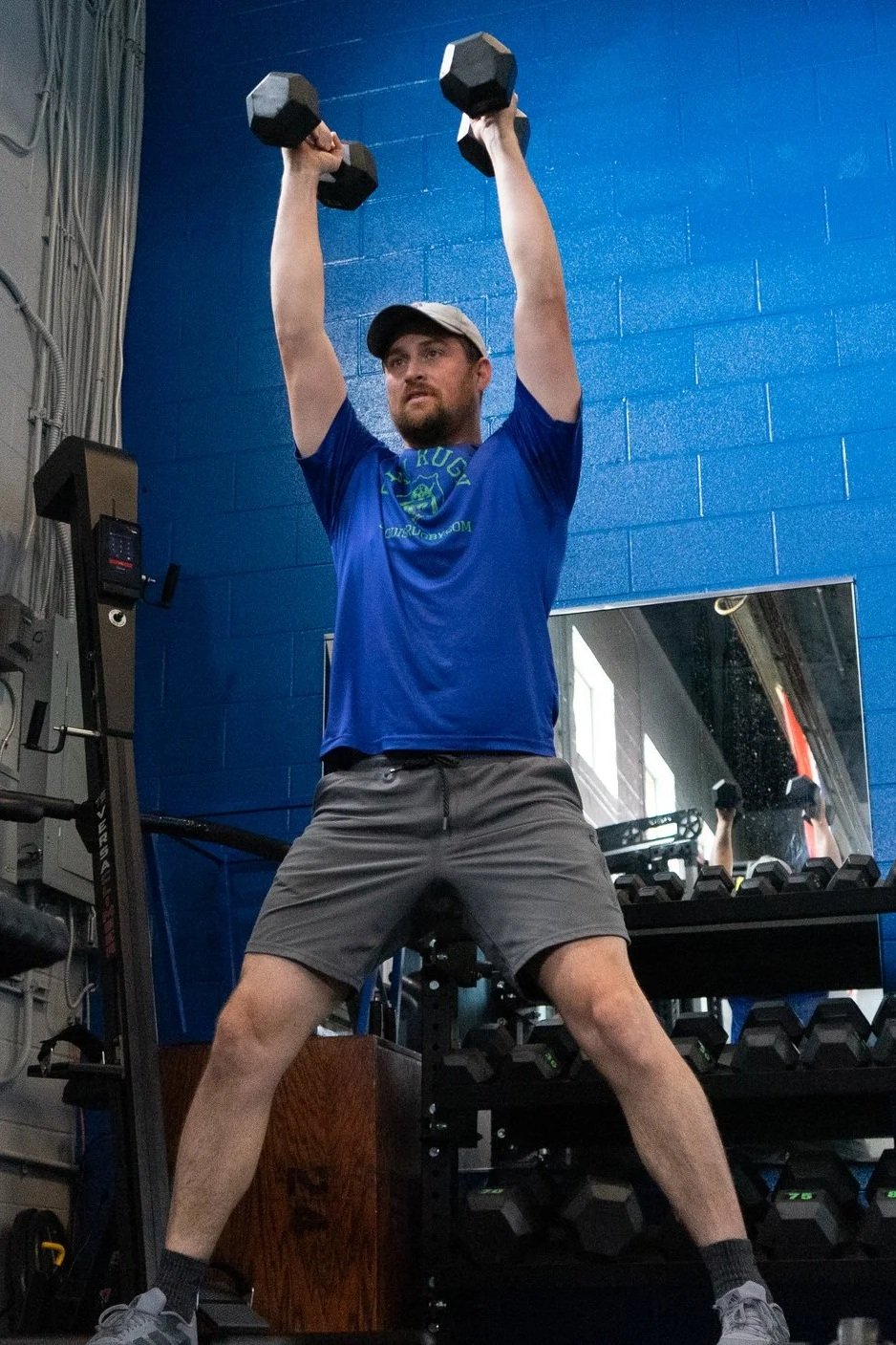 A man wearing a blue t-shirt, gray shorts, and a baseball cap is lifting two black dumbbells overhead in a gym. There are mirrors and workout equipment around him.
