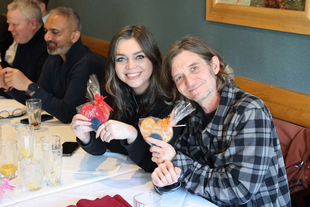 A group of people sitting at a table in a restaurant, smiling and holding decorated cookies; two people are in the foreground, a woman with dark hair and a man with long hair.