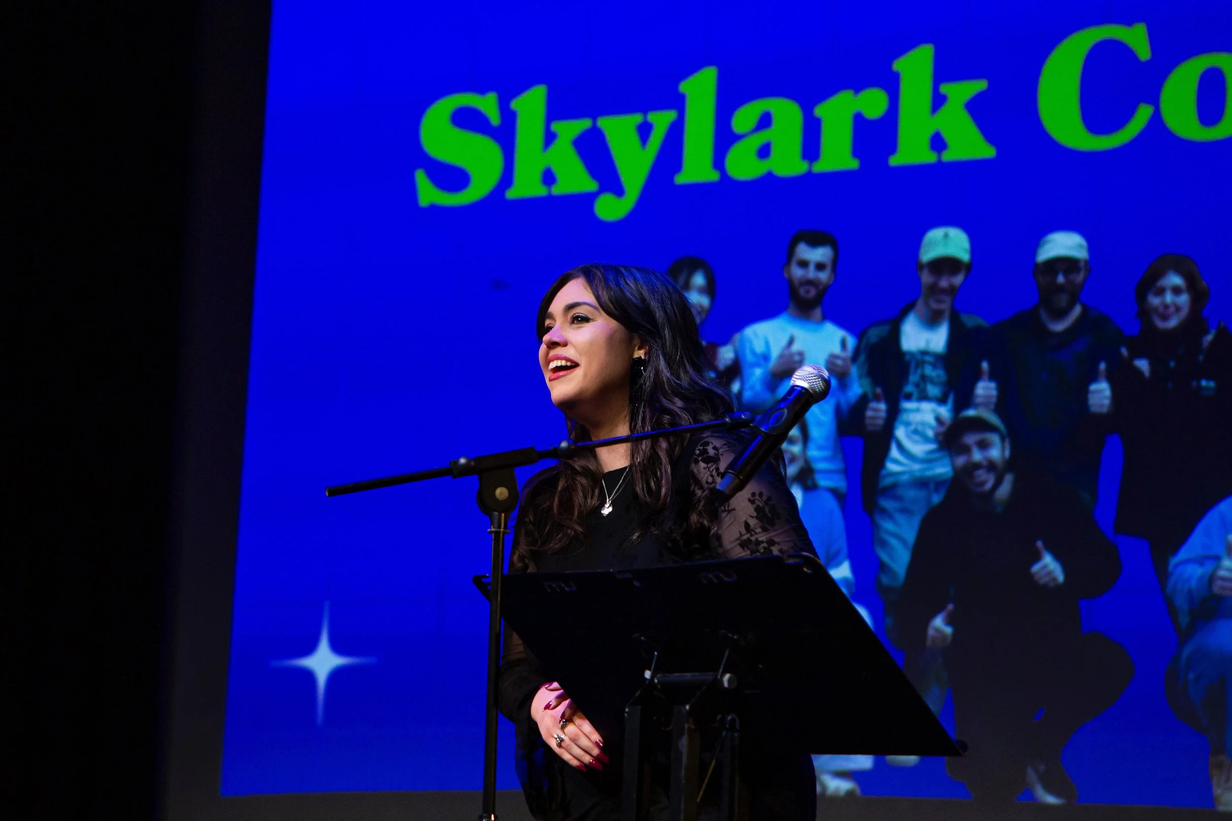 A woman singing or speaking at a microphone, with a large screen behind her displaying the word 'Skylark Co' and a group of people smiling and giving thumbs-up.