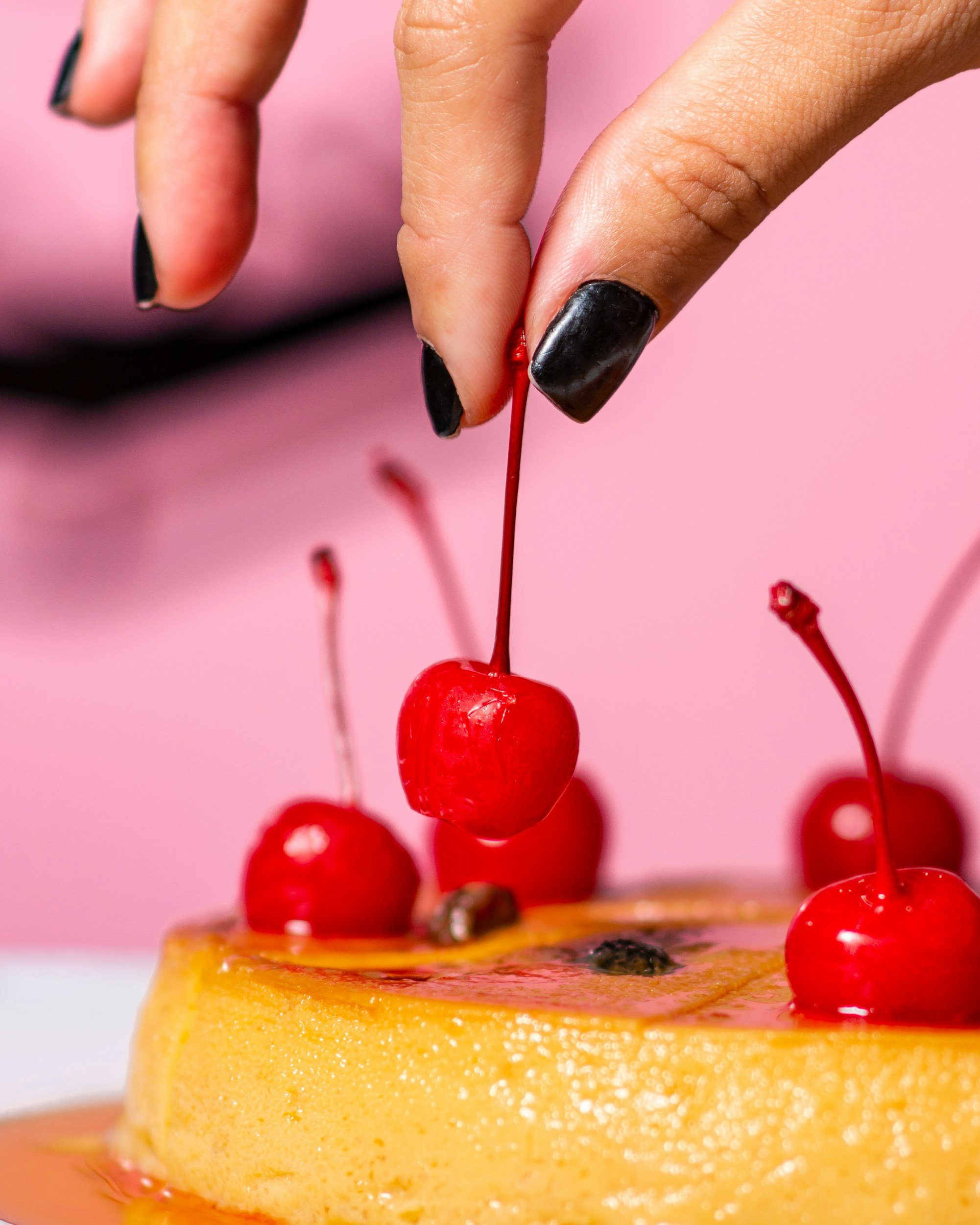 Close-up of a person’s hand with black nail polish, arranging maraschino cherries on a yellow dessert, possibly a cake, with a pink background.