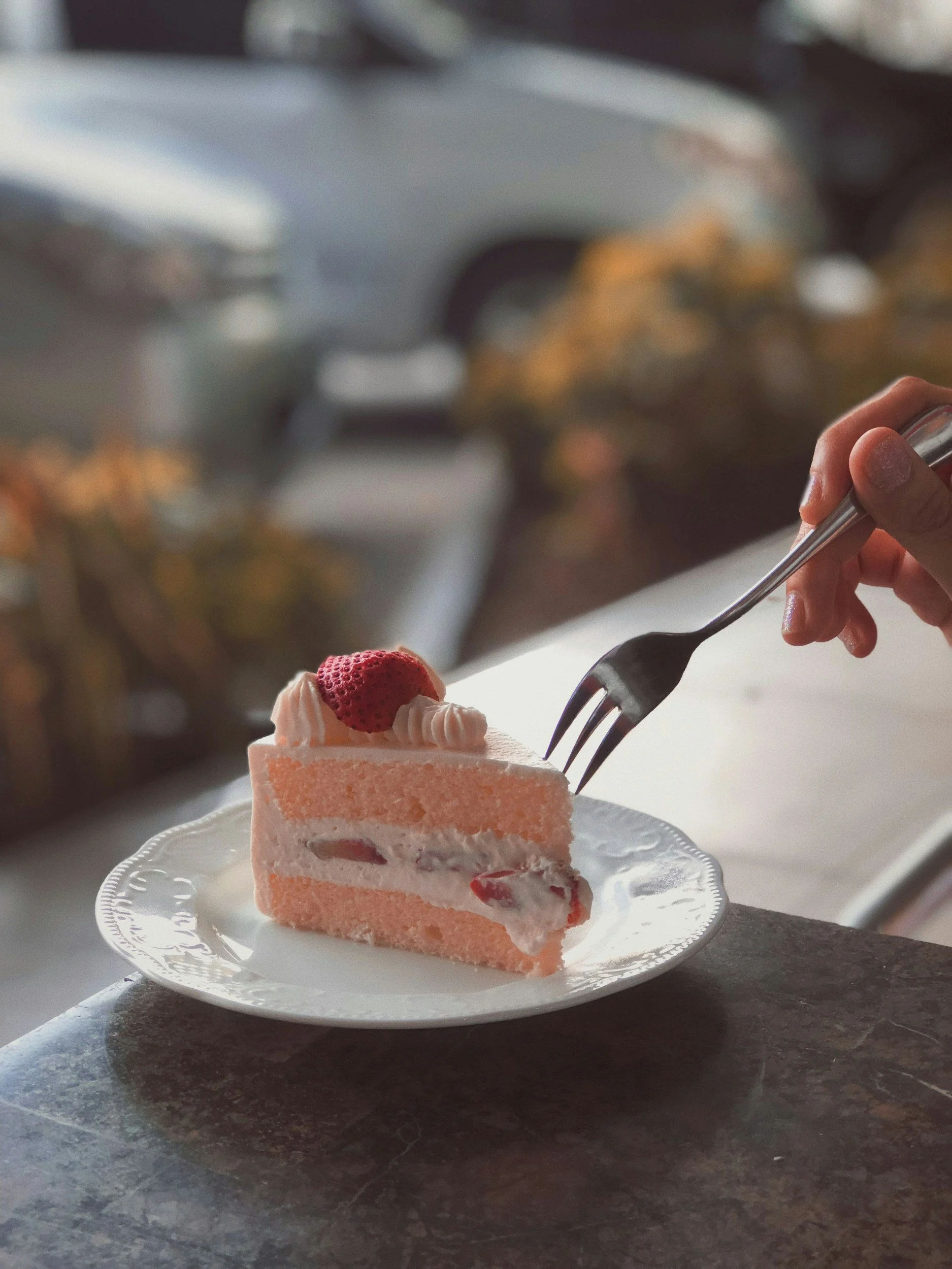 A slice of pink layered strawberry cake with whipped cream and a strawberry on top, on a decorative white plate.