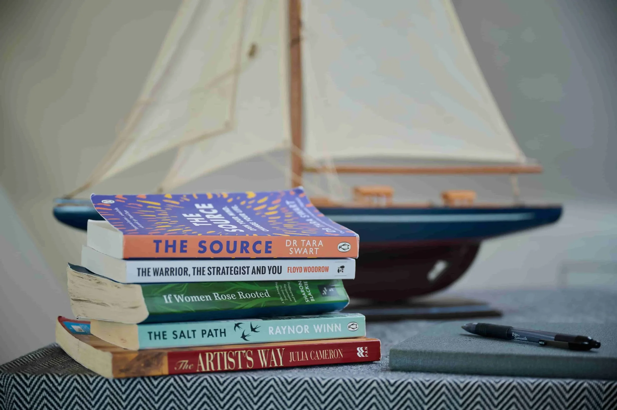 Stack of seven books on a table with a sailboat model in the background. The books from top to bottom are titled 'The Source' by Dr. Tara Swart, 'The Warrior, the Strategist and You' by Floyd Woodrow, 'If Women Rose Rooted' by Sharon Blackie, 'The Salt Path' by Raynor Winn, 'Addicted' by Julia Cameron, and an unnamed book at the bottom. A black marker pen is also on the table.