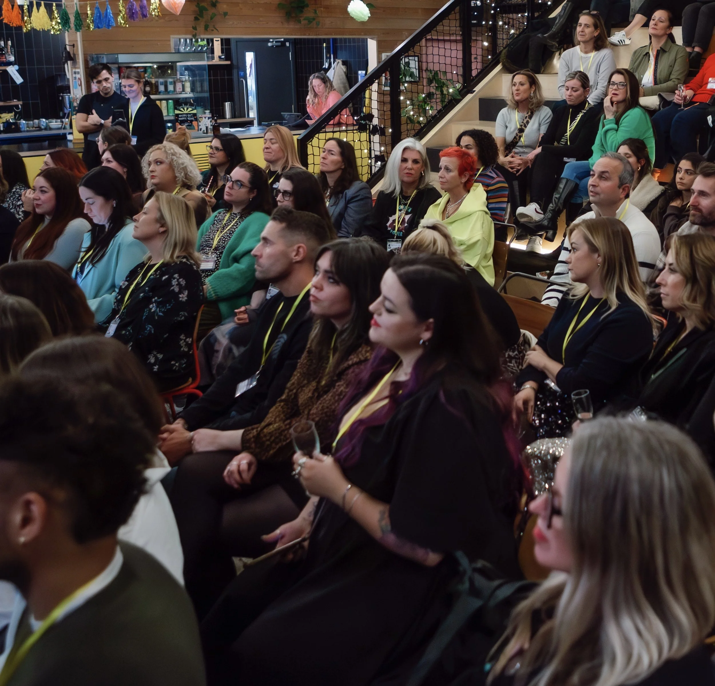 Audience attending a presentation or event at an indoor venue, seated on stairs and chairs, decorated with colorful banners and lights.