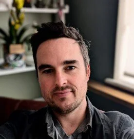 A man with dark hair and beard, wearing a dark shirt, sitting indoors near a window with a bookshelf behind him.