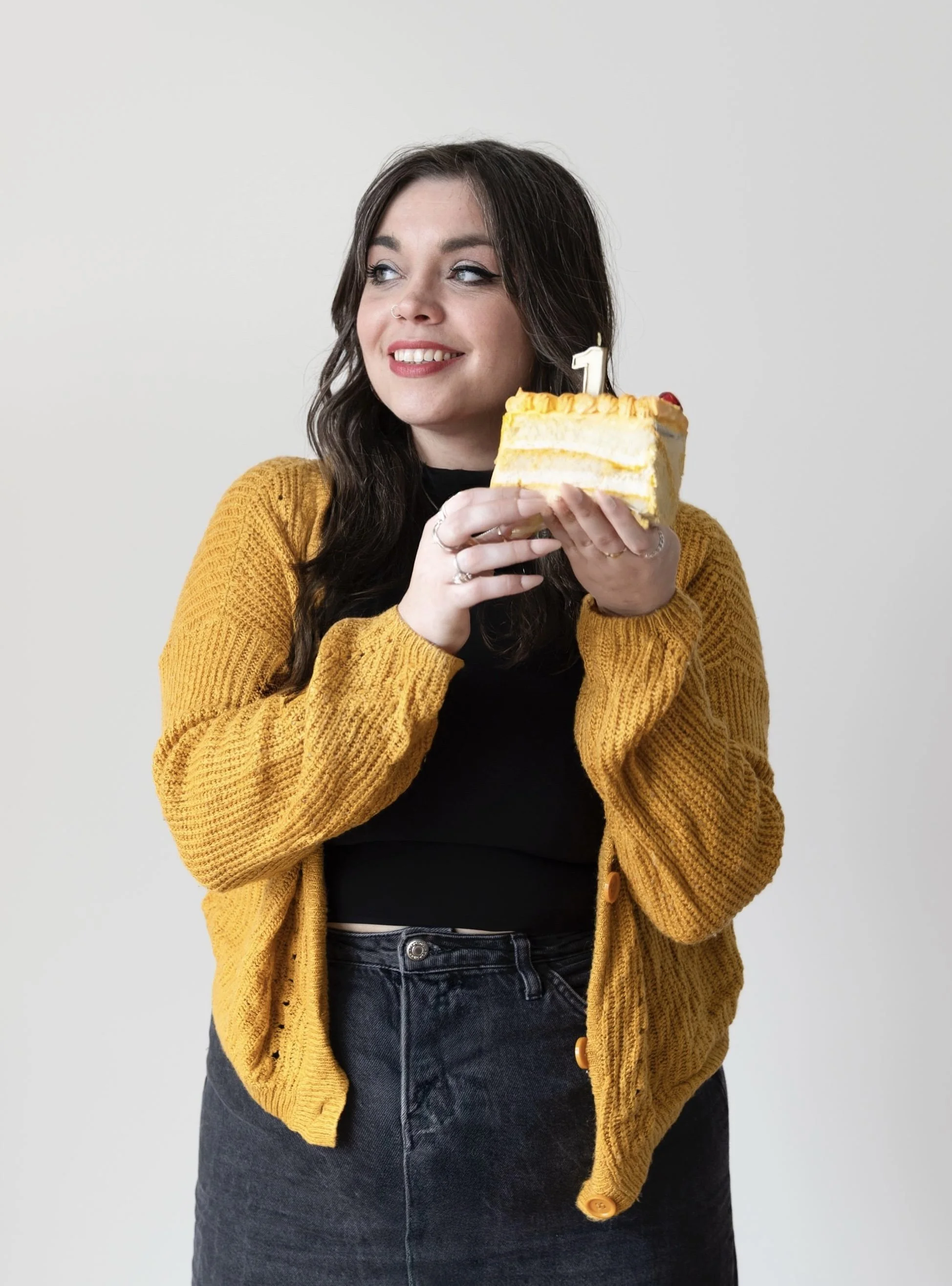 A woman with dark brown hair, wearing a mustard yellow sweater and black jeans, is smiling and holding a slice of layered cake with yellow frosting and a lit number one birthday candle.