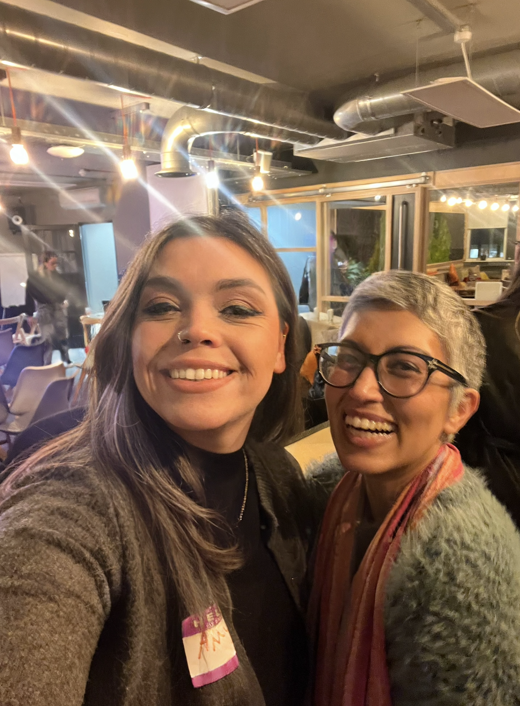 Two women smiling at a social gathering indoors with warm lighting, exposed ductwork, and a cozy, modern atmosphere.