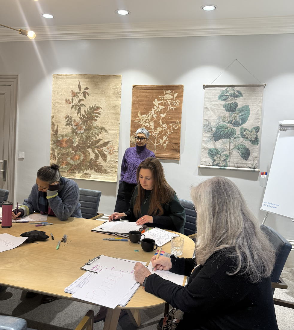 Four women are in a meeting room with artwork on the wall, papers and pens on the table, and a whiteboard in the background.