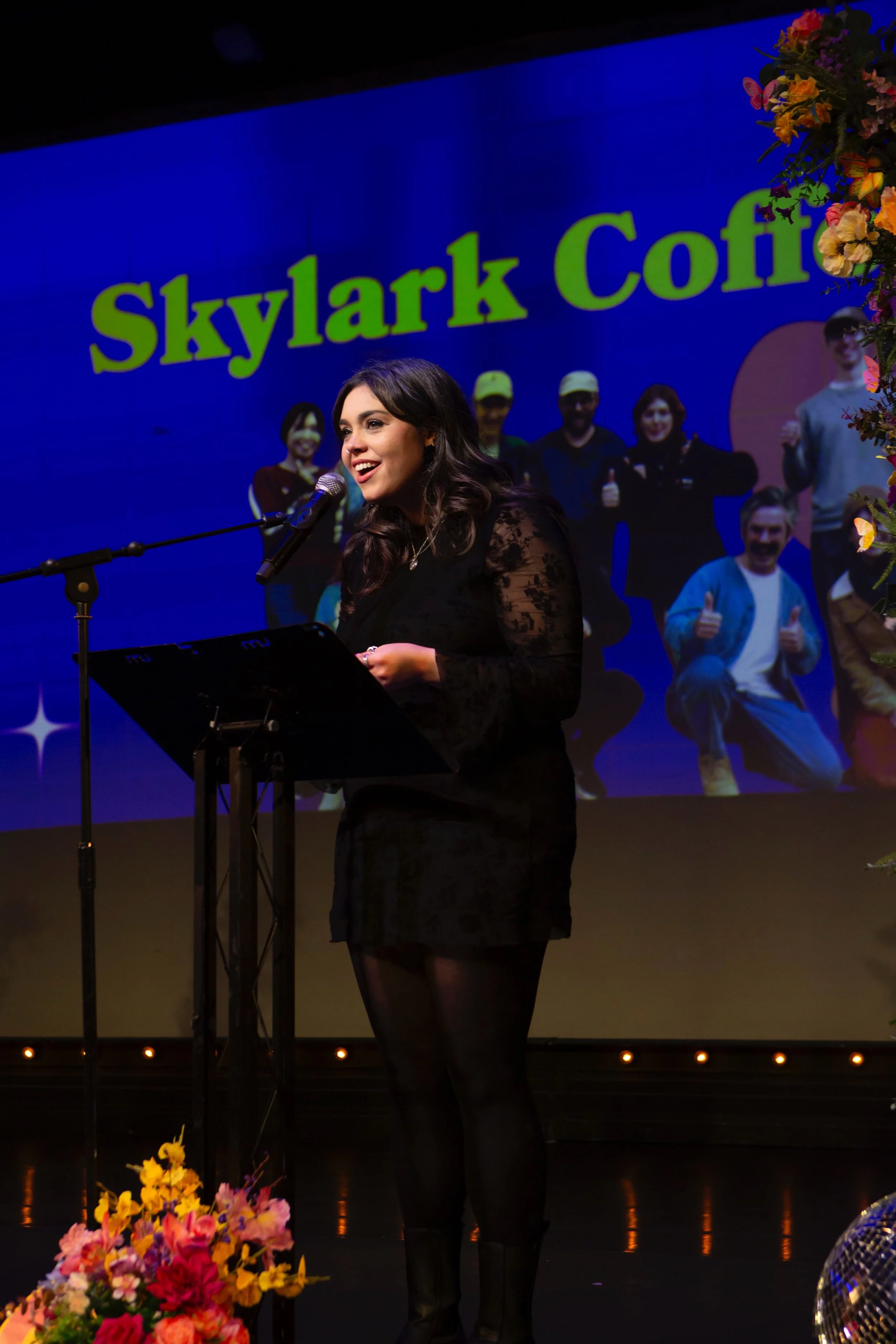 A woman standing at a microphone on stage, with a large screen behind her displaying the words 'Skylark Cofti' and a group photo. The stage is decorated with flowers and a disco ball.
