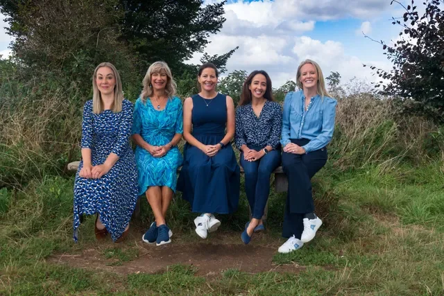 Five women sitting on a bench outdoors in a grassy area, smiling and dressed in blue clothing, with trees and a cloudy sky in the background.