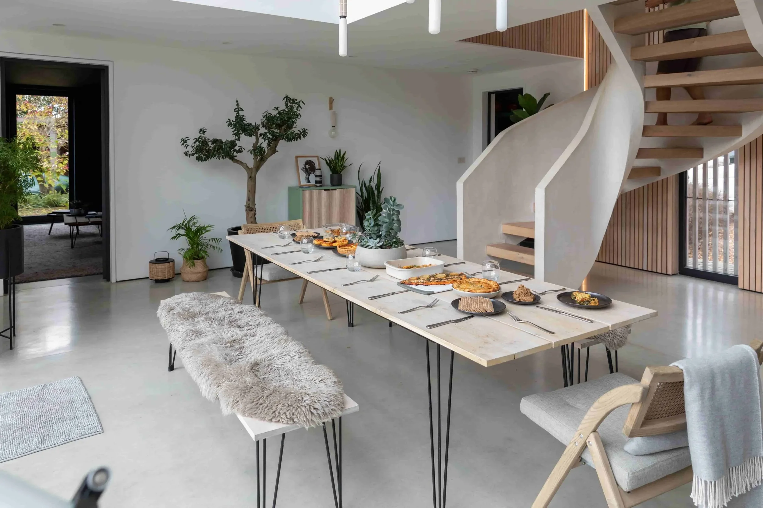 Contemporary dining room with a long table set for a meal, surrounded by different chairs and benches, with potted plants and trees adding greenery, and a staircase leading to an upper level.