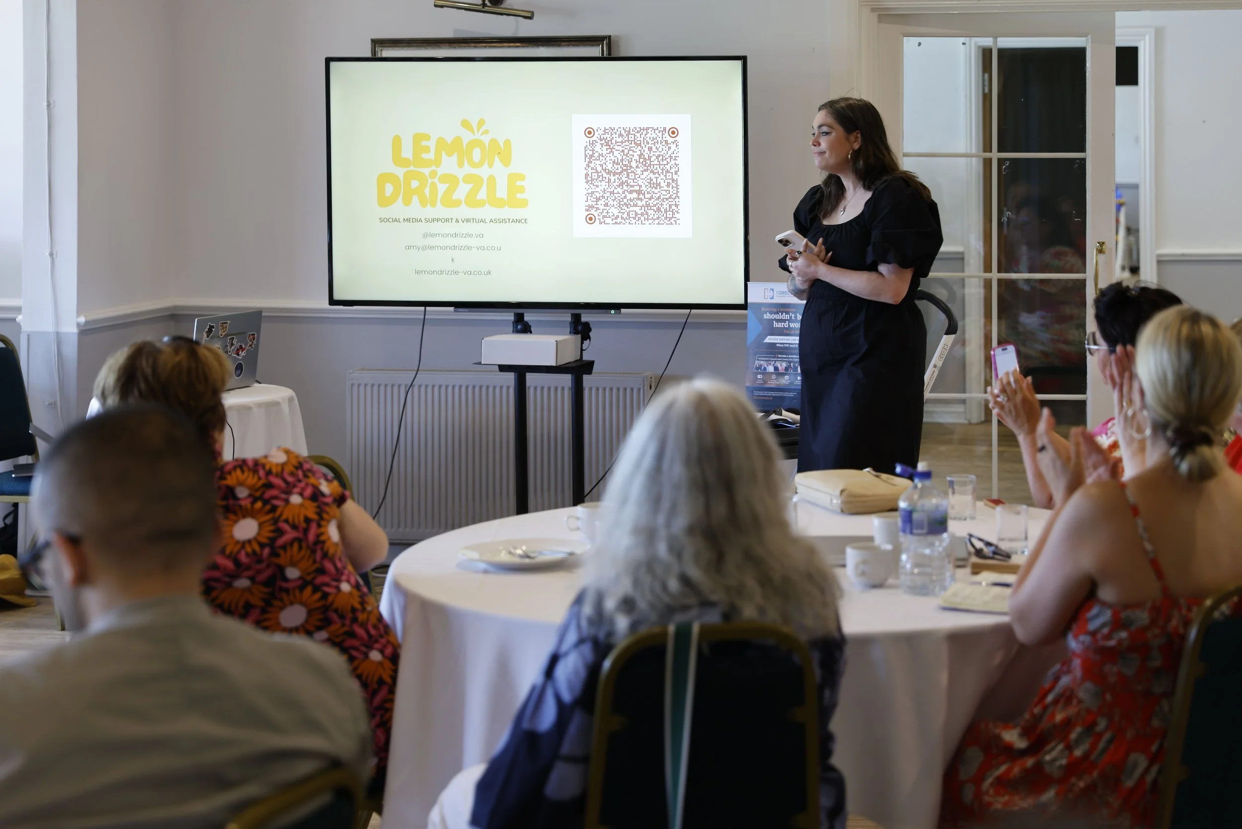 A woman presenting at a conference or workshop with a large screen behind her displaying a logo and QR code. Audience members are seated at tables, some taking notes or photos.