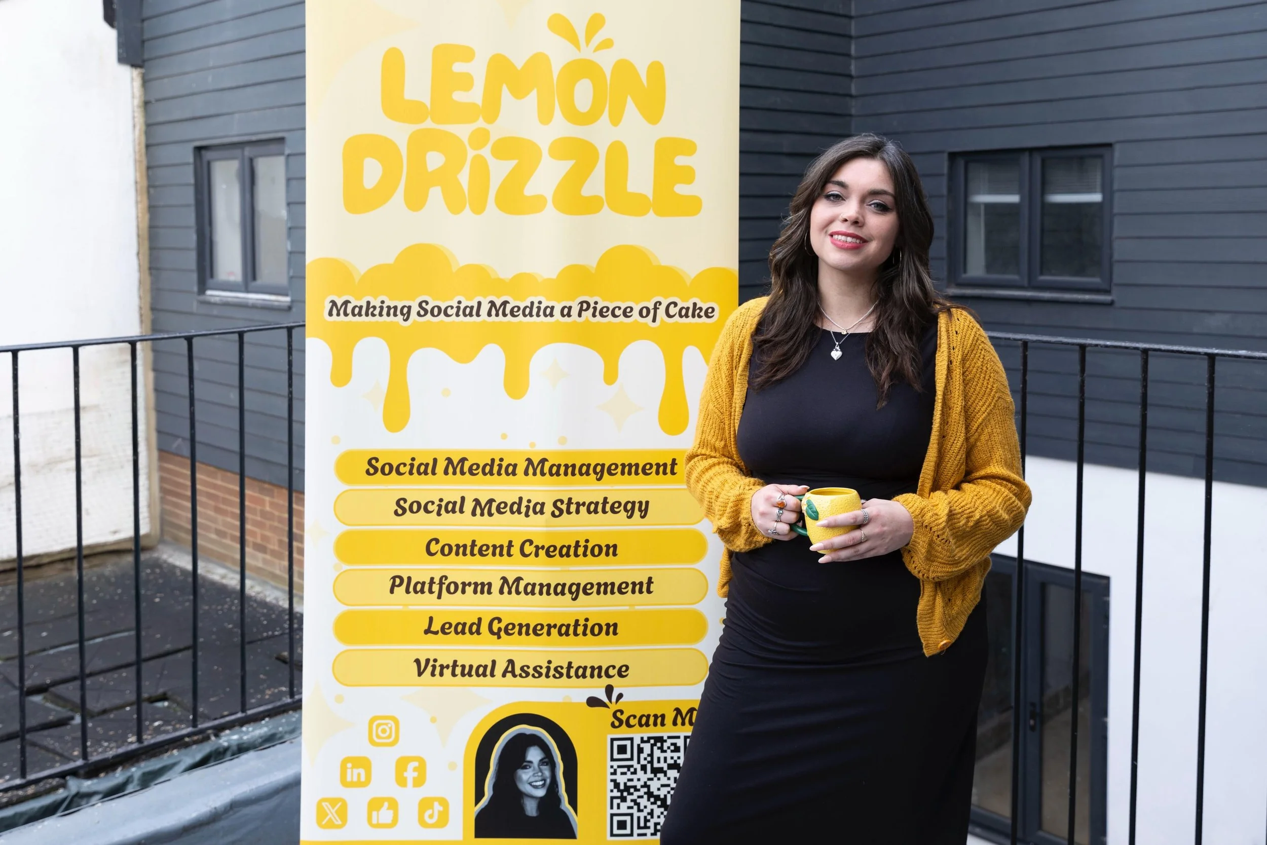 A woman with wavy brown hair and a yellow cardigan holding a yellow mug standing next to a large yellow sign with black text. The sign reads 'Lemon Drizzle' at the top and lists social media management services underneath, with icons for social media platforms and a QR code at the bottom. She is standing outside near a black fence and dark gray building with multiple windows.