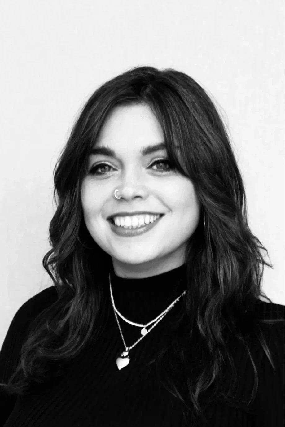 Black and white portrait of a woman smiling, wearing layered necklaces, with a nose ring, and wavy hair, against a plain background.