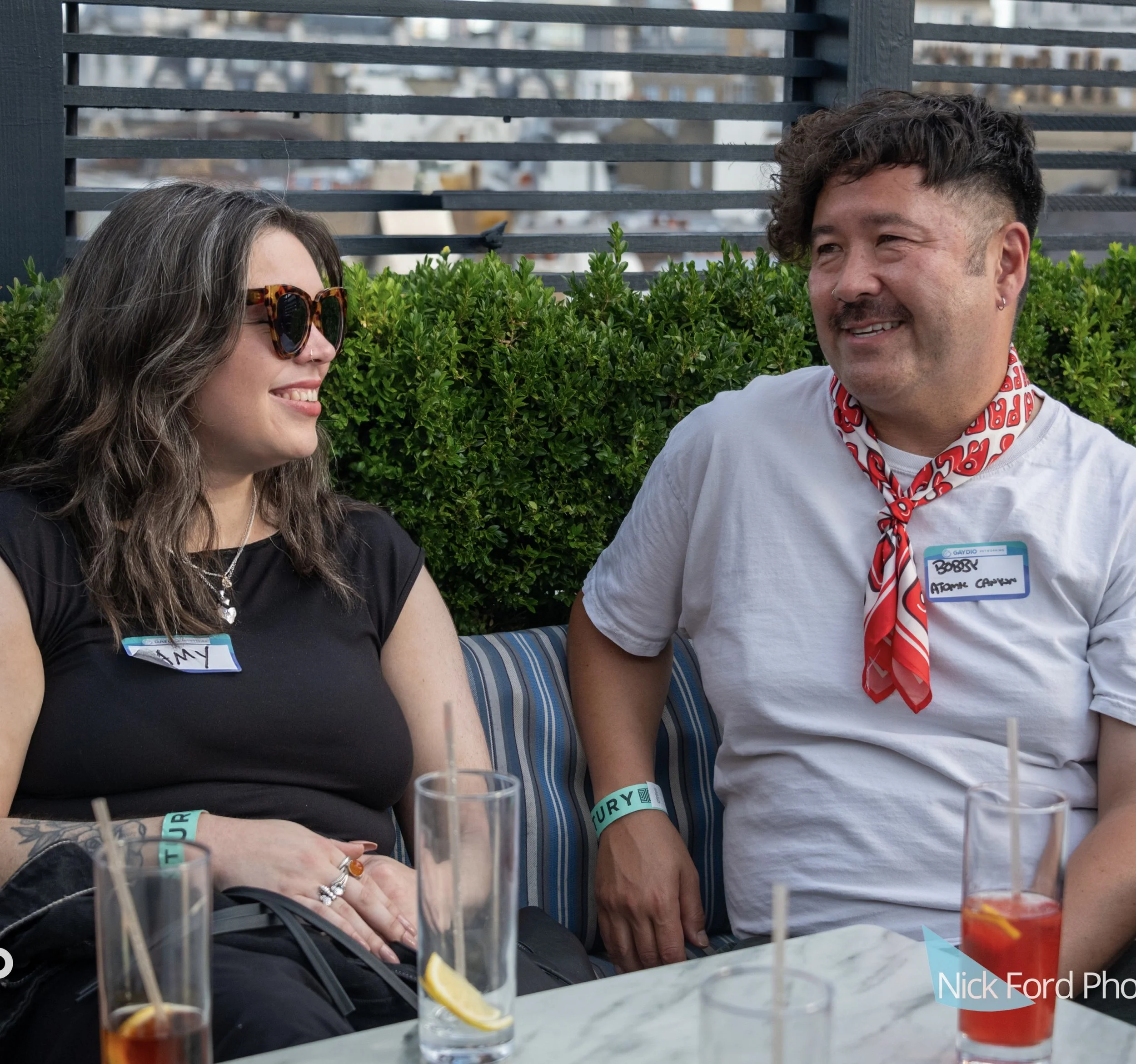 Two women sitting at a table outdoors, smiling and talking. One woman has long dark hair, wearing sunglasses and a black shirt, with a name tag that reads "Amy." The other woman has short dark hair, earrings, and a scarf around her neck, with a name tag that partially reads "Bobby" and the last name "Atome." There are drinks with lemon slices and straws on the table, and a green hedge behind them.