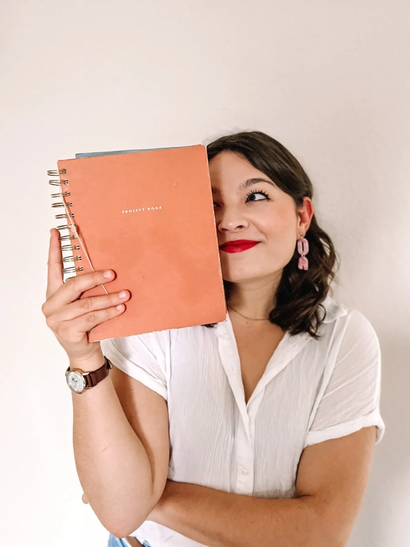 A woman with dark wavy hair, red lipstick, and pink earrings holds a peach-colored spiral-bound notebook labeled 'PROJECT BOOK' in front of her face, with a playful expression against a plain white background.