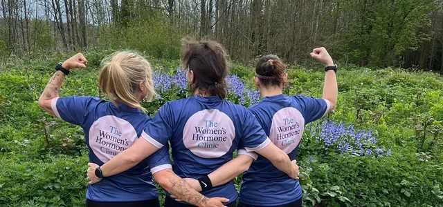Three women in matching navy T-shirts from The Women's Hormone Clinic, standing outdoors in a green and purple floral field, flexing their biceps with their backs to the camera.