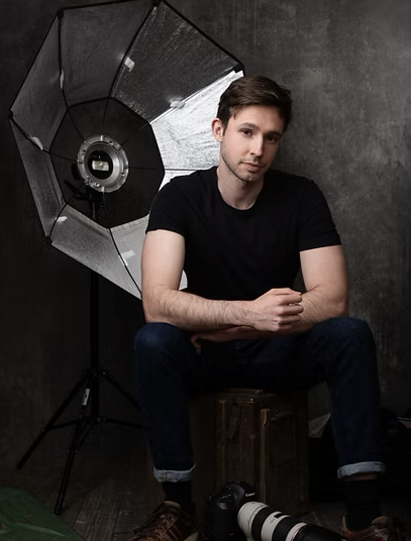 A young man with brown hair sitting on a wooden box in front of photographic studio equipment, including a large reflector, with a dark textured background.