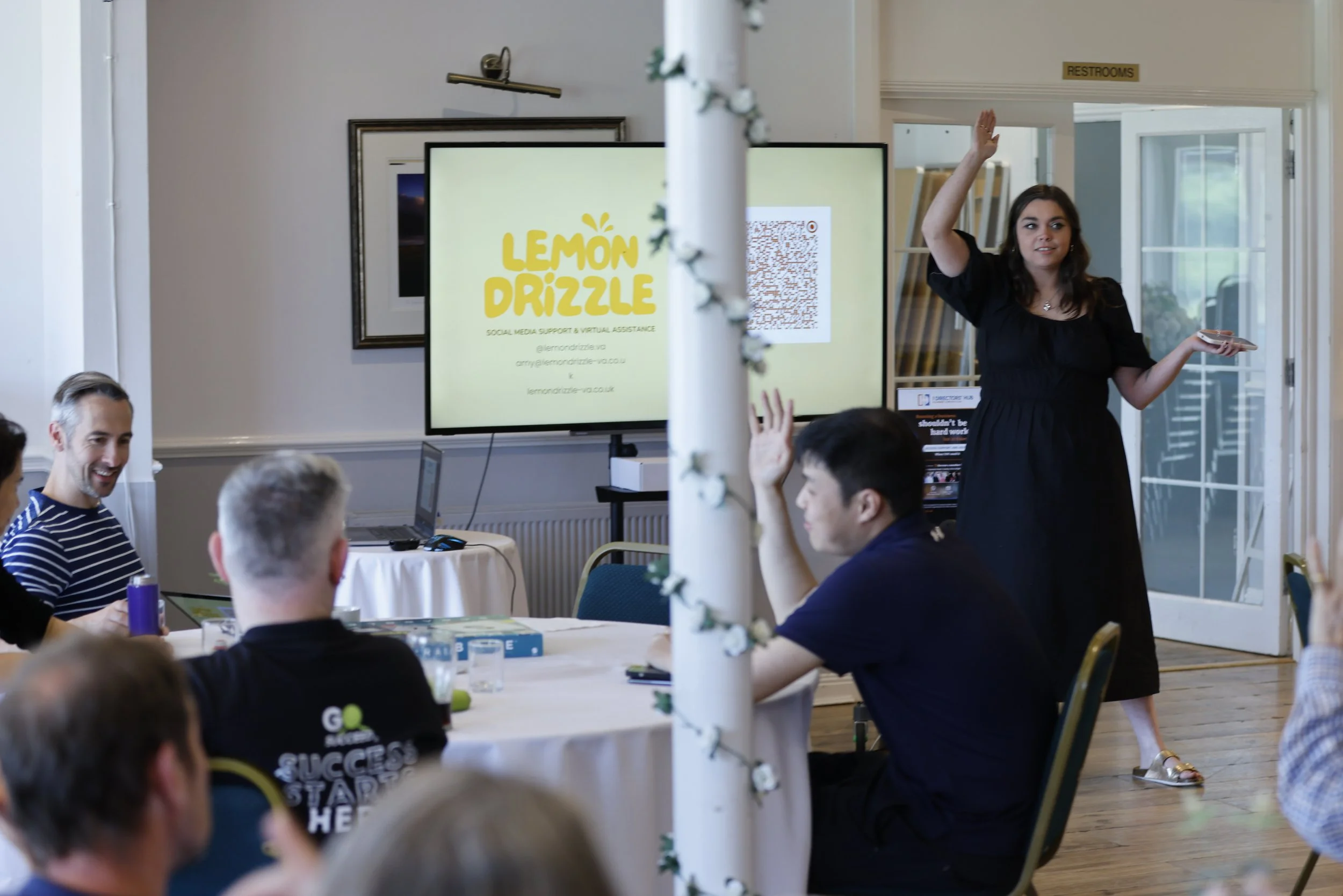 A woman in black dress standing and speaking to a group of people at a meeting or workshop, with a presentation slide reading 'Lemon Drizzle' visible in the background.