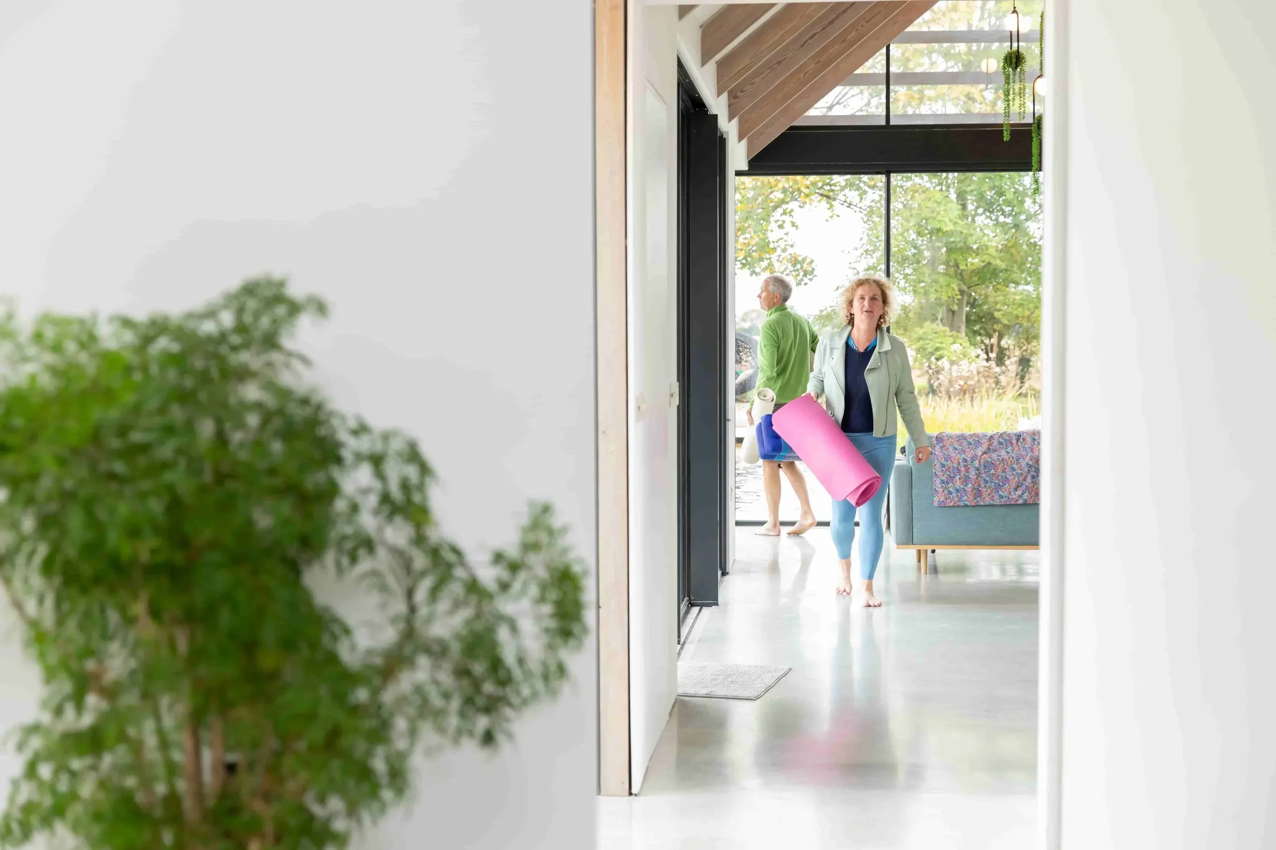 Woman walking through a home with yoga mat and water bottle, smiling, with a man standing outside near a tree in the background.