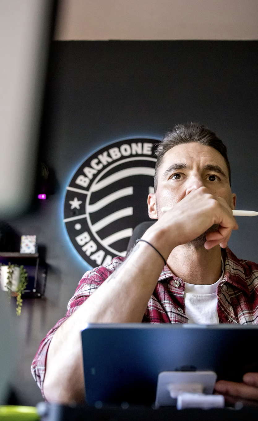 young man sitting at a desk in a modern workspace, holding a pen near his face, with a logo on the wall behind him reading 'Backbone' and 'BRAND' with a circular design.