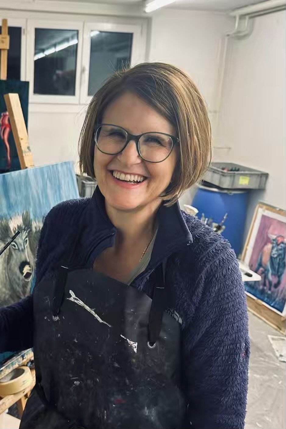 Eine lachende Frau mit Brille vor einer Malerei, umgeben von Kunstwerken und Malutensilien in einem Atelier.