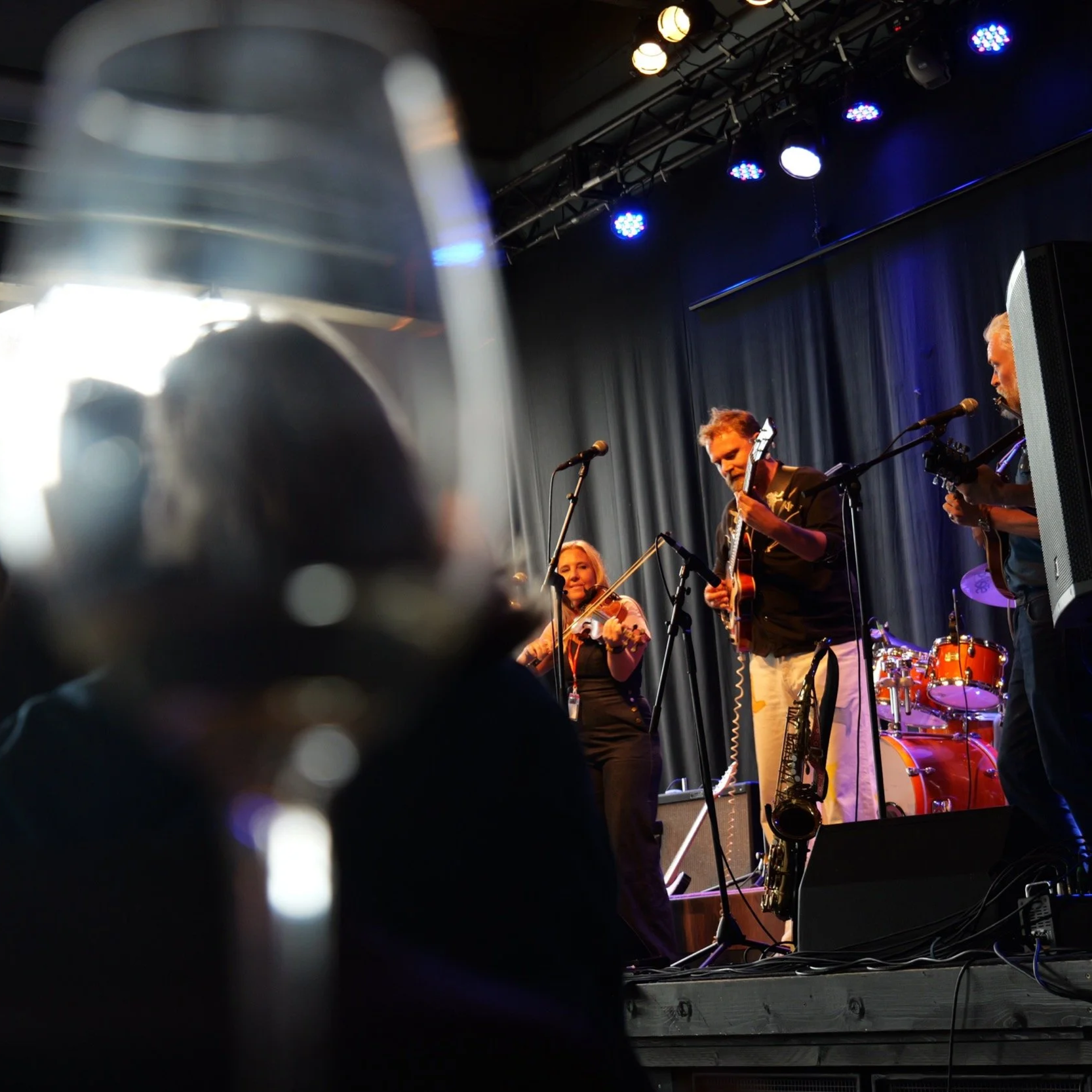 En grupp musiker som spelar på scen, inklusive en man med elguitar och en kvinna med fiol, under en konsert med scenerbelysning.