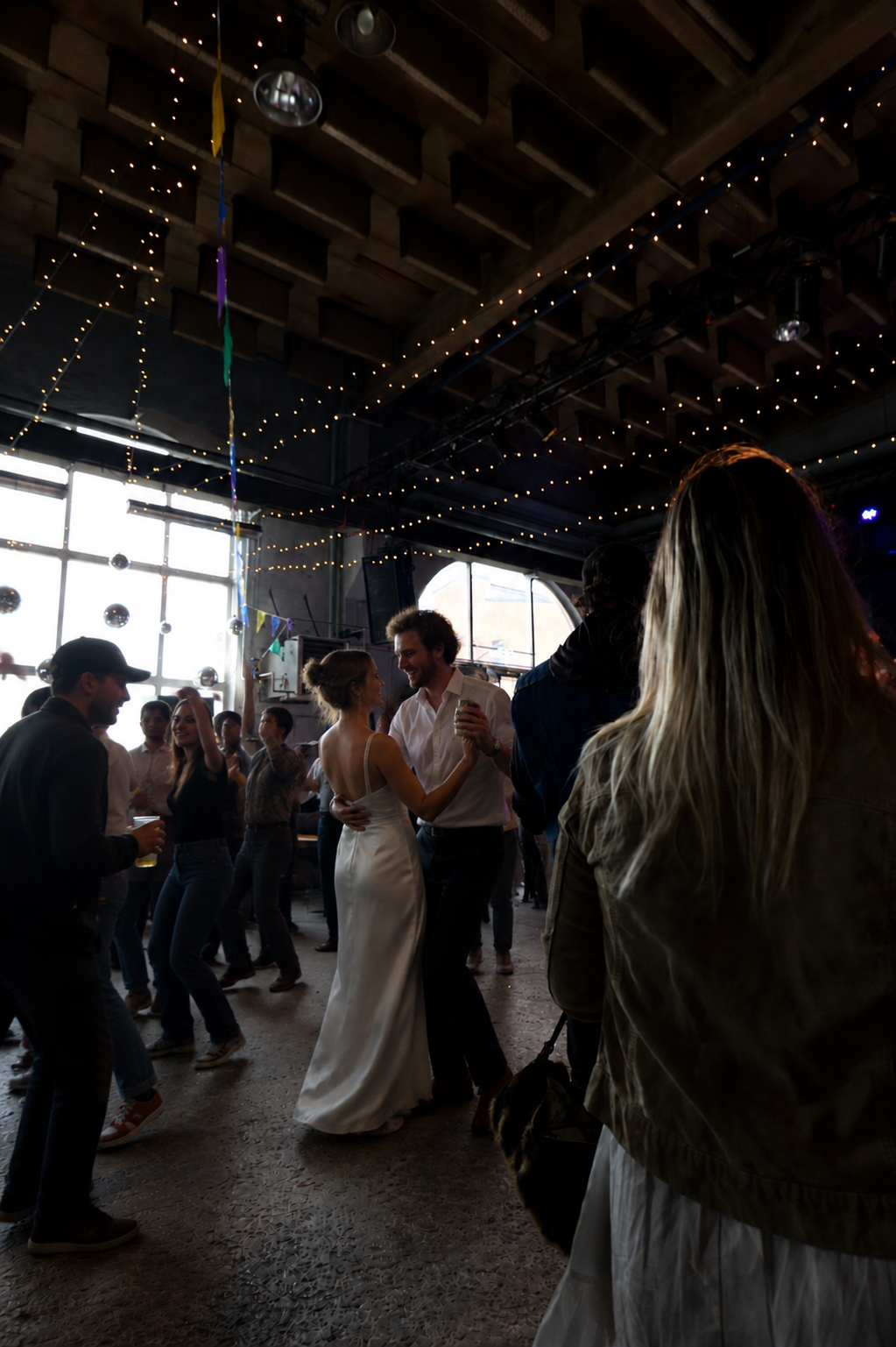 Folk dansar och njuter på ett festligt inomhusevent, dekorerat med strålkastare, string lights och ballonger.