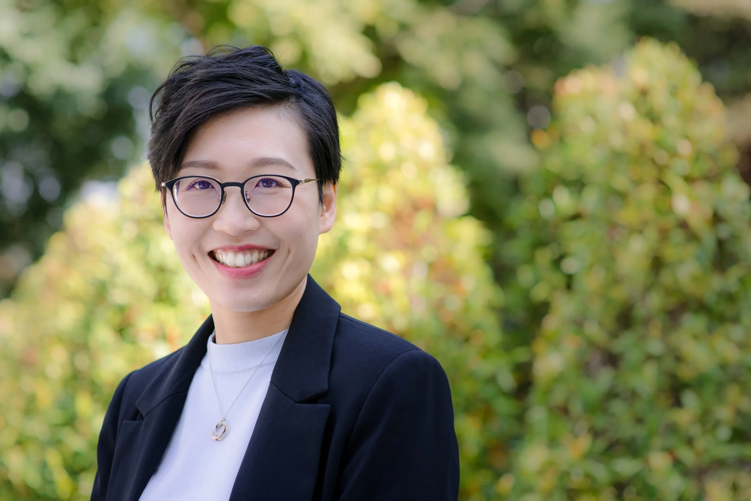 A woman with short black hair wearing glasses, a dark blazer, and a white shirt, smiling outdoors with a blurred green and yellow foliage background.