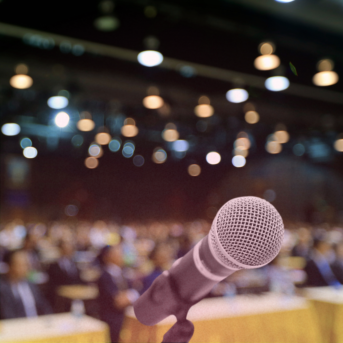 Microphone in the foreground with an audience and blurred lights in the background, indicating a conference or presentation setting.
