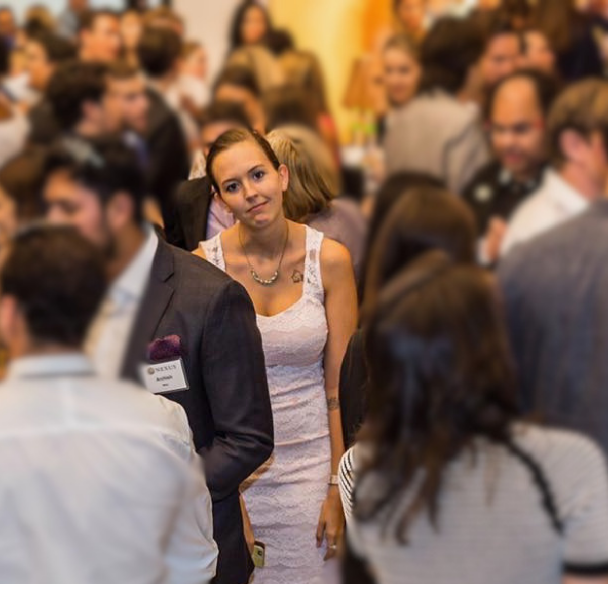 Woman in a white lace dress standing in a crowded room with many people, some wearing suits, at a social or professional event.