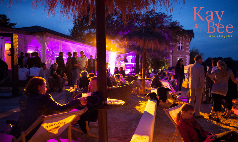 People socializing at an outdoor event during evening with colorful lighting, some sitting on lounge chairs, others standing and talking, with a stage and purple lighting in the background.