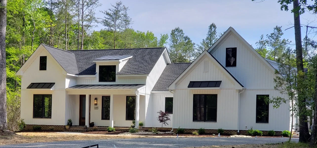 Modern white farmhouse-style house with black window frames and a shingled roof, surrounded by trees and a gravel driveway.