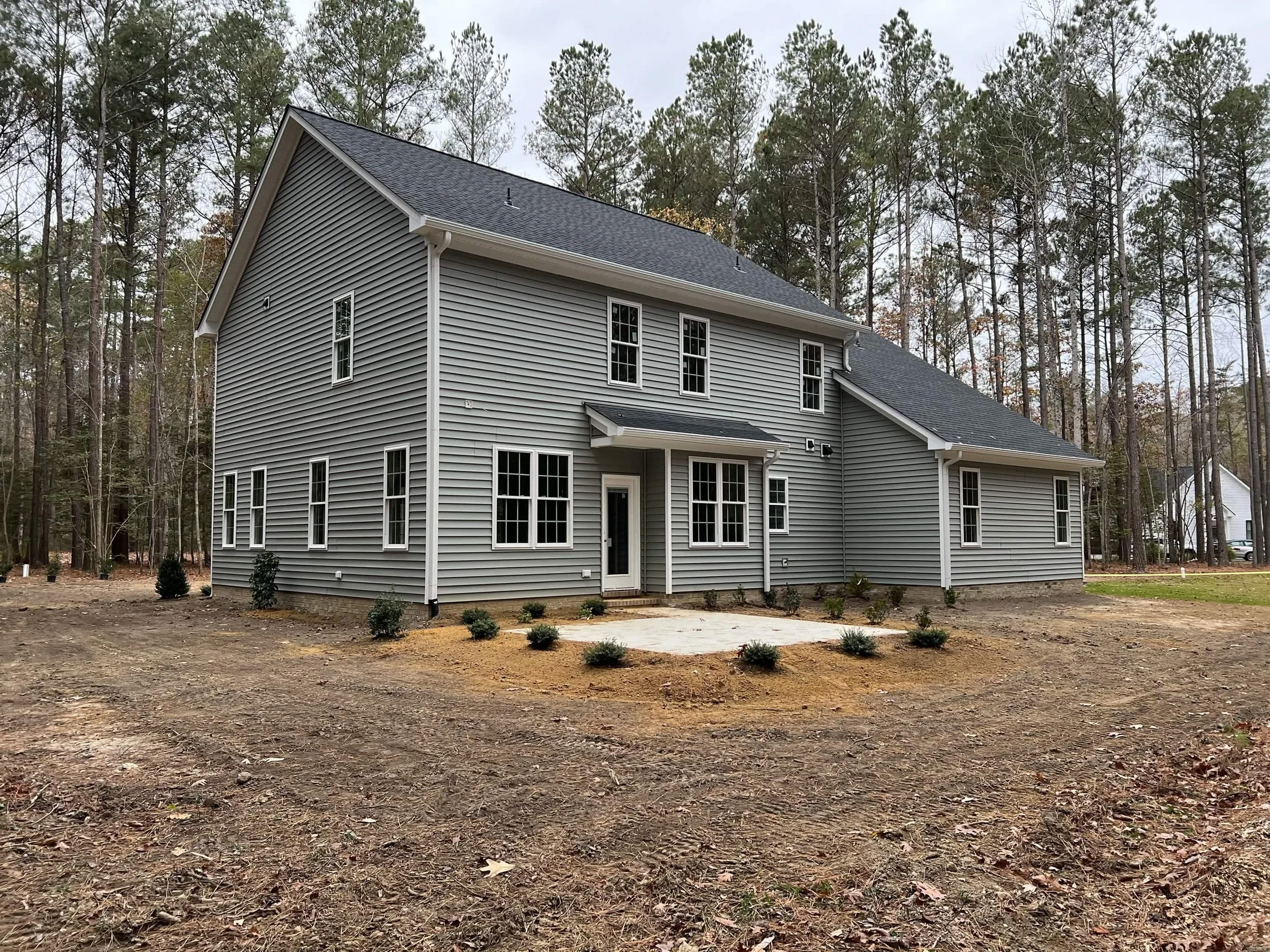New gray two-story house with multiple windows, surrounded by a bare yard and some small bushes, with a wooded background.