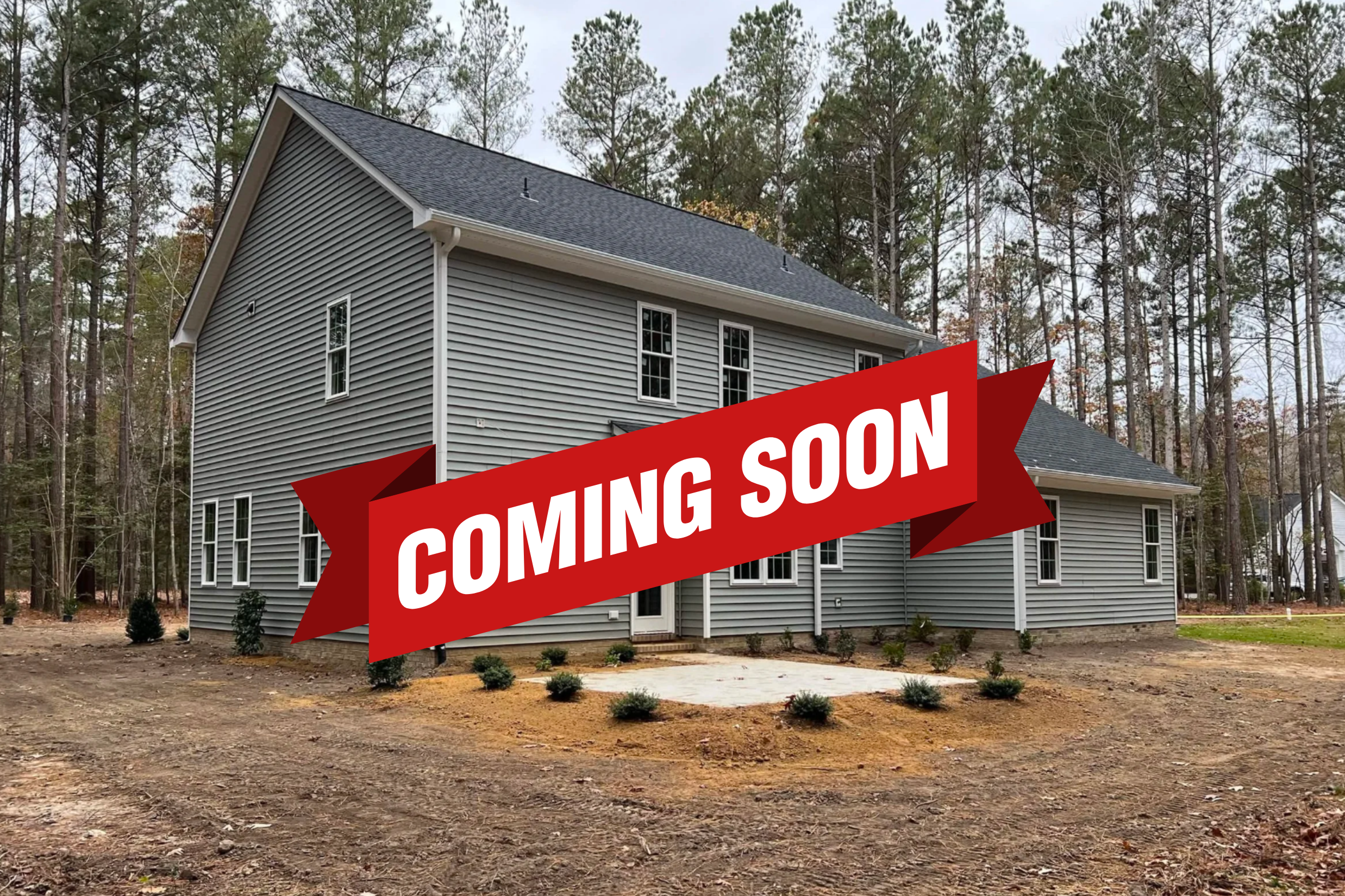 New gray two-story house with multiple windows, surrounded by a bare yard and some small bushes, with a wooded background.