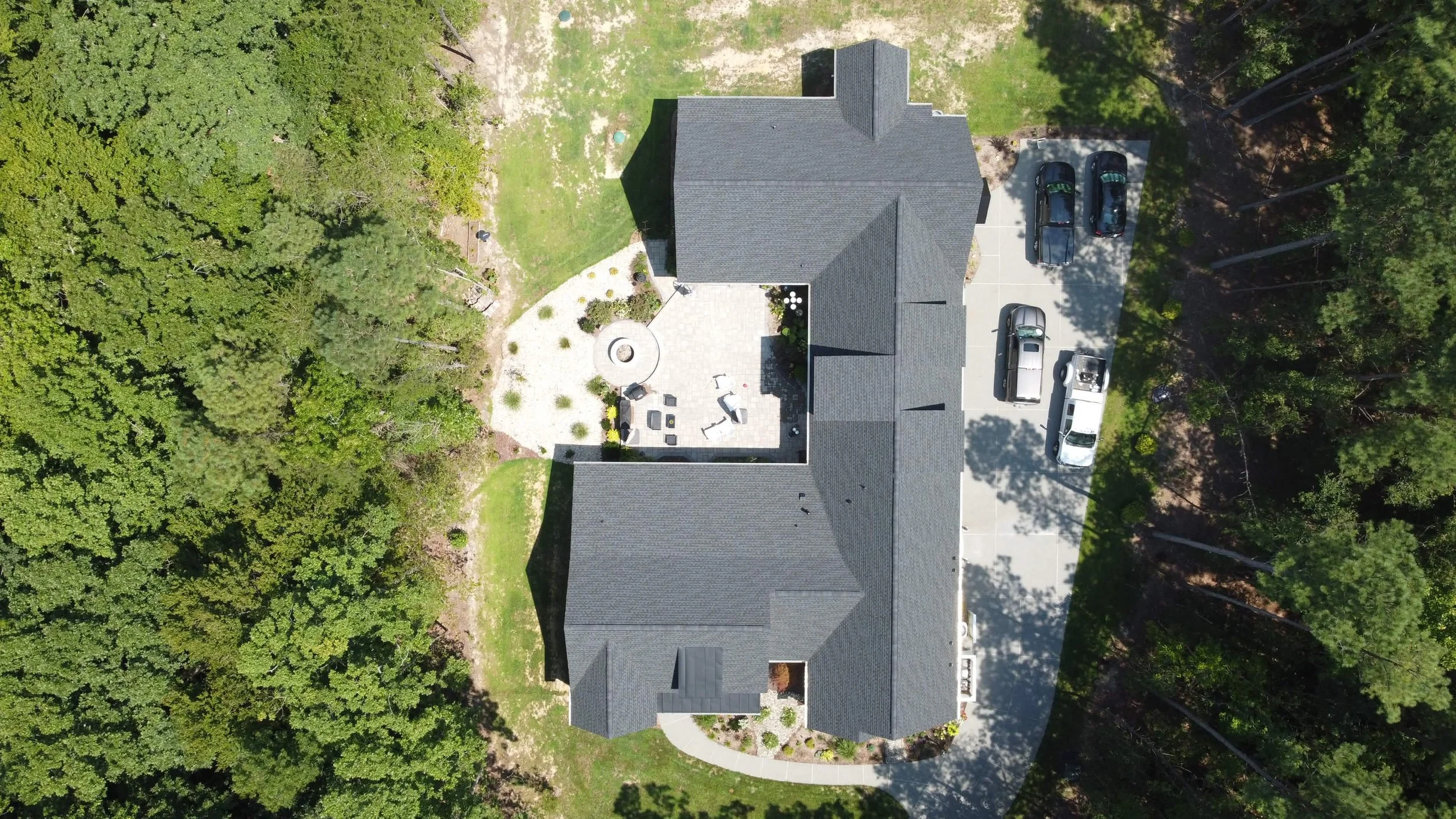 Aerial view of a house surrounded by trees, with a driveway and parked cars. The backyard has a patio with outdoor furniture and a fire pit.