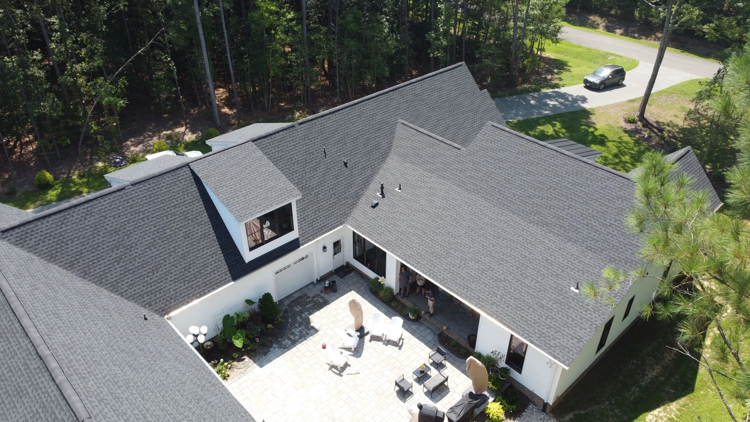 Aerial view of a suburban house with gray tiled roof, white exterior walls, and a spacious backyard patio with outdoor furniture, surrounded by tall trees and a driveway with parked cars.