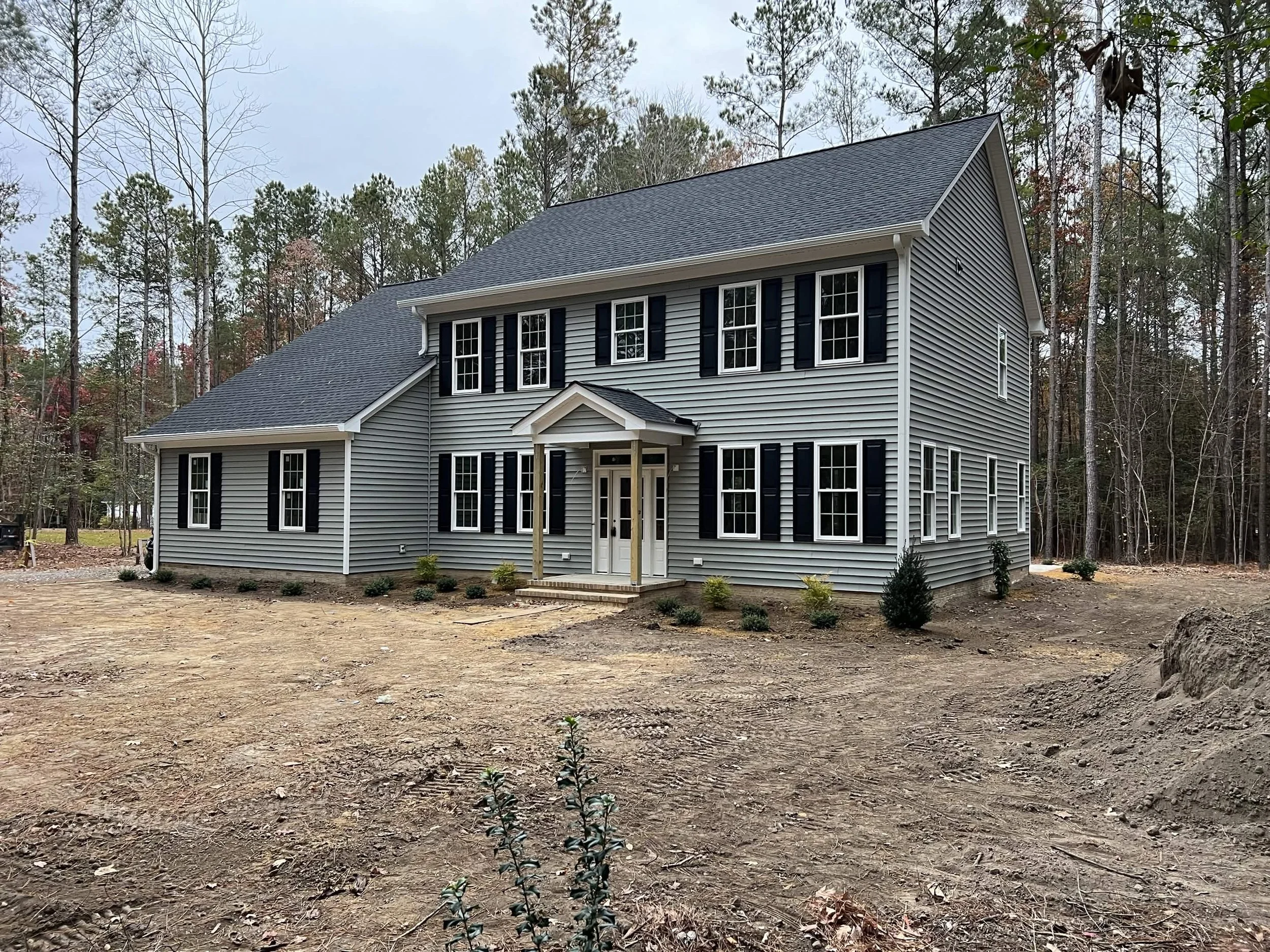 Newly built two-story house with gray siding and black shutters, surrounded by a cleared yard and forested area in the background.
